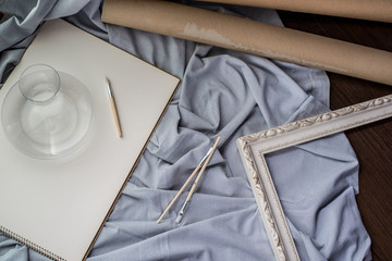 View of a creative space: brushes, vintage frame and blank paper on blue fabric.