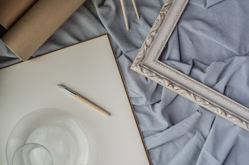 View of a creative space: brushes, vintage frame and blank paper on blue fabric.