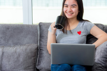Asian woman holding a coffee cup smile and looking during a work break, Working remotely with a laptop on the sofa in the house, Cute girl teens happy use internet computer to communication from home