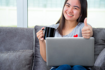 Asian woman holding a coffee cup smile looking and show thumb in work, Working remotely with a laptop on the sofa in the house, Cute girl teens happy use internet computer to communication from home