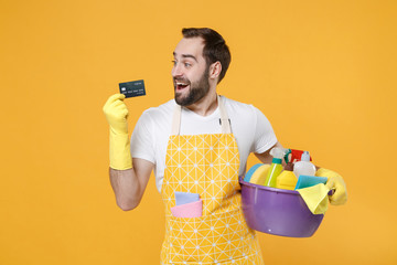 Excited young man househusband in apron rubber gloves hold basin with detergent bottles washing...