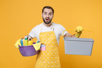 Puzzled young man househusband in apron rubber gloves hold basin detergent bottles washing...