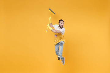 Fototapeta na wymiar Crazy screaming young man househusband in apron rubber gloves hold broom while doing housework isolated on yellow wall background studio portrait. Housekeeping concept. Mock up copy space. Jumping.