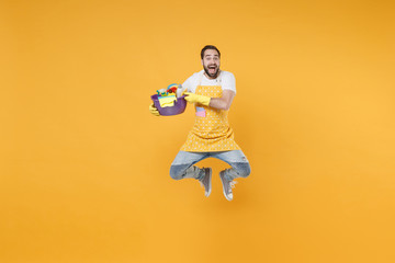 Fototapeta na wymiar Excited man househusband in apron gloves hold basin detergent bottles washing cleansers doing housework isolated on yellow background. Housekeeping concept. Mock up copy space. Jumping, having fun.