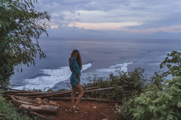 Naklejka premium A beautiful girl stands on a cliff overlooking the waves of the blue ocean. Tropical paradise and exotic nature on the island of Bali. Model with different emotions of happiness and joy.