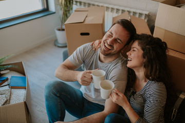 Top view photo of young happy couple drinking coffee while cardboard boxes are all around them. Young couple is moving in to their new home. - Powered by Adobe