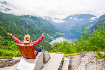 Female tourist sitting on the Queen Sony Chair at the Flydalsjuvet Viewpoint. The Geiranger village...