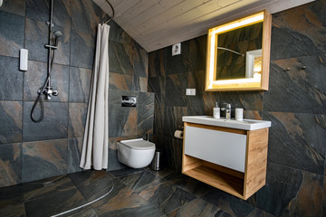 Interior of modern stylish bathroom with black tiled walls, curtain shower place and wooden furniture with wash basin and big illuminated mirror.
