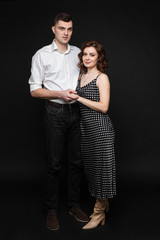 Handsome man with short black hair in white t-shirt and black trousers hugs with his wife with short chestnut wavy hair in long grey dress
