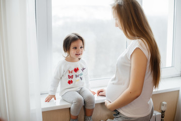 A pregnant mother is standing near the window with her little daughter