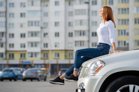 Young Stylish Redhead Woman In White Sweater, Fashionable Blue Denim Jeans Pants And Black Sneakers Sitting On A Hood Of New Expensive Car On City Street Enjoying Warm Summer Day.