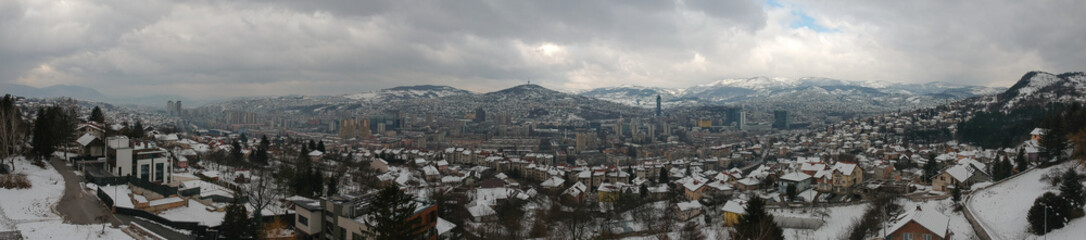 Fototapeta na wymiar Winter drone panorama of Sarajevo, capital of Bosnia and Herzegovina, taken on a snowy winter day with clouds. Snow is seen on the houses and the mountains in the back.