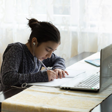 Young Girl Sitting At The Dining Table With Laptop At Home Schooling, Online Virtual Classroom Video Conference, Distant Education. Active Participation At The Lesson With Earphones, Doing Homework.