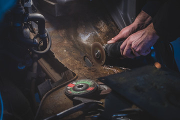 Bare hands of a man using an angle grinder to cut off the excess rusty metal from a car body in the...
