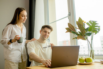 beautiful caucasian married couple together at home, man work from home, at freelance. careful wife support and hug him from back. man use laptop, surf the Net