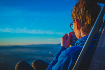 Rear view of a young woman with glasses enjoying a cup of coffee on the lookout point at the top of the hill at sunset. Female hiker drinking coffee at the summit.