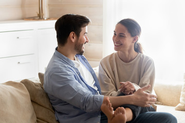 Happy young couple resting on comfortable sofa, chatting at home. Smiling mixed race girl putting legs on boyfriend, relaxing on couch in living room, enjoying sincere conversation, dating concept.