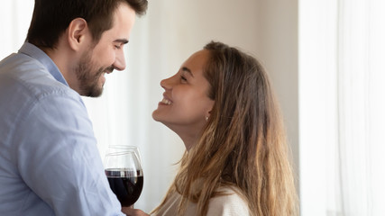 Emotional loving young family couple looking into eyes, toasting drinking wine indoors, side view. Close up head shot affectionate spouses clinking glasses, celebrating anniversary or dating at home.
