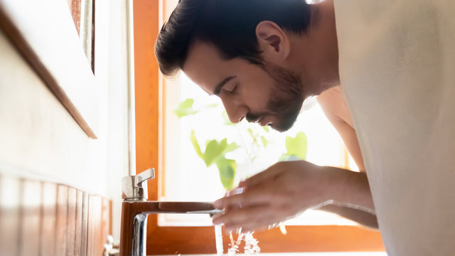 Side View Young Bearded Man Washing Face With Running Tap Water, Bend Over Sink In Bathroom. Millennial Handsome Guy Doing Morning Skincare Hygiene Routine After Showering, Head Shot Close Up View.