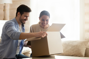 Happy millennial couple sitting on cozy sofa, unboxing cardboard parcel. Overjoyed young spouses clients consumers unpacking carton box, satisfied with fast delivery service, high quality purchase. - obrazy, fototapety, plakaty