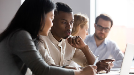 Multiracial serious colleagues sit at desk talk brainstorm using computer cooperate at office briefing, serious diverse coworkers speak discuss business ideas at boardroom meeting together