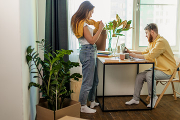young caucasian woman do household chores, unpacks boxes after moving into new house while her husband work with laptop at home, man at freelance. married couple in new apartment