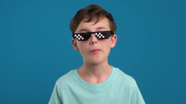 Portrait happy boy chewing gum and learning to inflate bubbles and bursting in pixel sunglasses it joyfully smiling looking at camera slow motion on blue background. Childhood. Emotions people