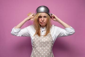 Fototapeta premium tired girl cook in kitchen clothes with a pan on her head on a colored background, woman housewife in stress