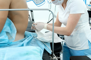 An anesthetist in white medical clothes, a mask and sterile gloves holds a needle in his hands. Near the back of the patient who will undergo epidural anesthesia. Preparing for a surgical operation.