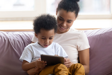 Happy african american mother sitting with son on couch looking at tablet device. Smiling diverse family engage in online shopping with boy together. Mom and chilld wathcing on smartphone.