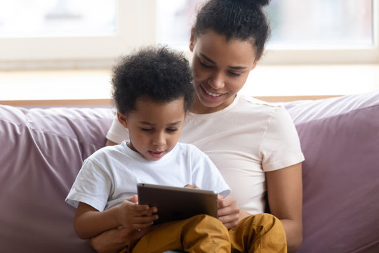 Happy African American Mother Sitting With Son On Couch Looking At Tablet Device. Smiling Diverse Family Engage In Online Shopping With Boy Together. Mom And Chilld Wathcing On Smartphone.