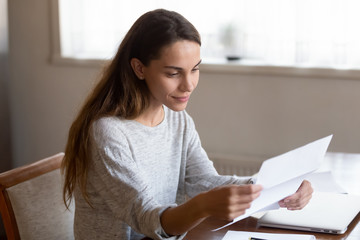 Happy focused young lady sitting alone at table, reading paper letter with good news. Smiling millennial girl woman received positive answer in pleasant postal correspondence, feeling optimistic.
