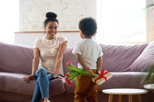 Rear view african american son holding flowers, bouquet and gift behind back, congratulate young diverse mother with mothers day or birthday, adorable child making surprise to mum at home.