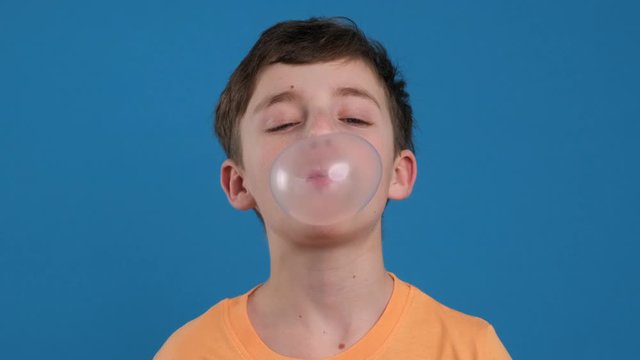 Happy boy chewing gum and learning to inflate bubbles and bursting it joyfully smiling looking at camera slow motion on blue background. Child. Childhood. Emotions of people. Life style