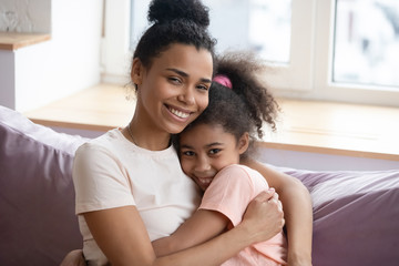 Close up headshot portrait of millennial african american mother and teen daughter looking at camera sitting on couch. Happy smiling diverse family embrace together in living room at home.