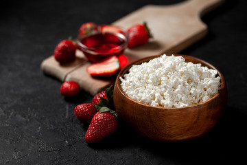 homemade cottage cheese in a wooden plate without additives lies with strawberries and red jam on a cutting board, healthy food on a black table