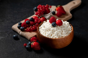 fresh cottage cheese with strawberries, raspberries, blueberries, currants in a wooden plate against a dark background, healthy milk food with wild berries on a black table
