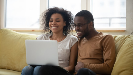 Happy african american family having good time using laptop. Young smiling husband and wife sitting on couch at home looking at computer screen and watching video.