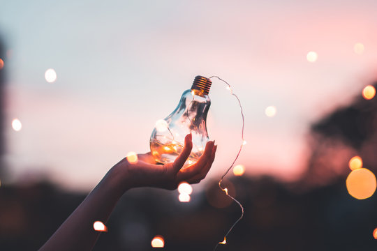 Close-up Of Woman Holding Illuminated Light Bulb Against Sky At Night