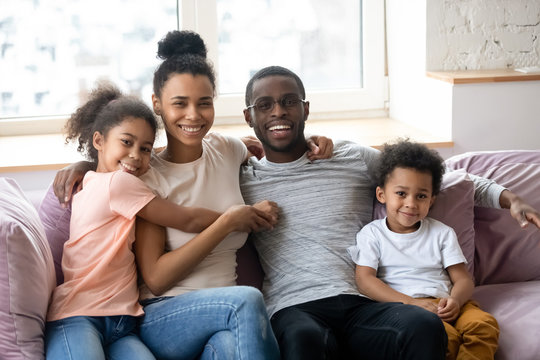 Portrait of african american happy family with two children looking at camera. Cute daughter and son with parents sit on couch in living room. Father and mother enjoying time together with children.
