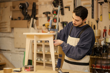 portrait of young caucasian carpenter making wooden chair in the factory, wearing uniform, man keen...