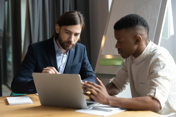Obraz premium Diverse multiracial male business colleagues sit at desk in boardroom brainstorm negotiate use laptop together, serious multiethnic coworkers talk discuss financial ideas work on computer in office