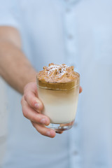 Cup of Dalgona or whipped coffee served on a glass and a wholemeal orange cake behind. Wooden black background