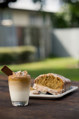 Cup of Dalgona or whipped coffee served on a glass with a bar of chocolate and a wholemeal cake. Outdoor background
