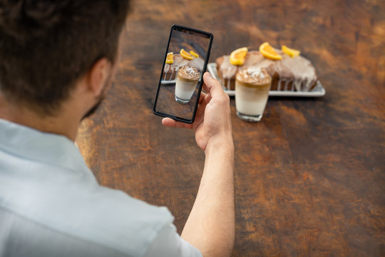 Man taking a photo with mobile at a glass of Dalgona coffee and an orange cake on wooden background