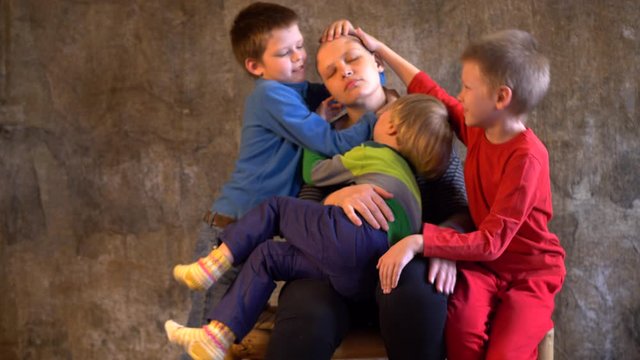 Attractive girl with shaved bald head. Beautiful middle-aged woman with cancer without hair. Children in colorful clothes pat their mother’s head and regret after chemotherapy session