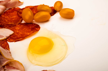 A broken chicken egg without a shell, slices of sausage and bacon and tomatoes on a white background. Close up.