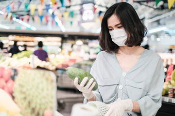 Asian woman wearing protect face mask and rubber gloves shopping food, fruit and vegetable in grocery department store. Girl choosing mango in supermarket during coronavirus crisis, covid19 outbreak.