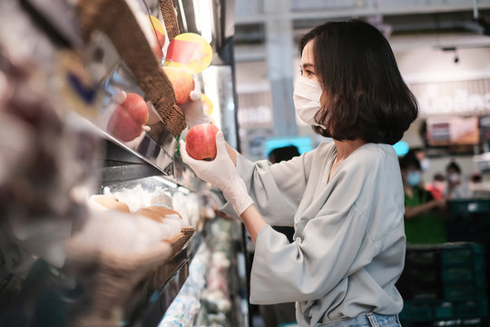 Asian woman wearing protect face mask and rubber gloves shopping food, fruit and vegetable in grocery department store. Girl choosing apple in supermarket during coronavirus crisis, covid19 outbreak.