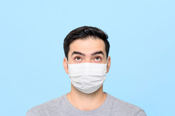 Young man wearing medical face mask thinking and looking up to empty space above isolated on light blue studio background